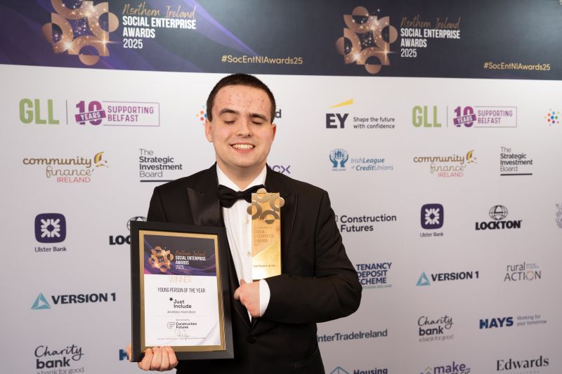 Andrew Standing against a sponsor wall in black tie smiling with his award.
